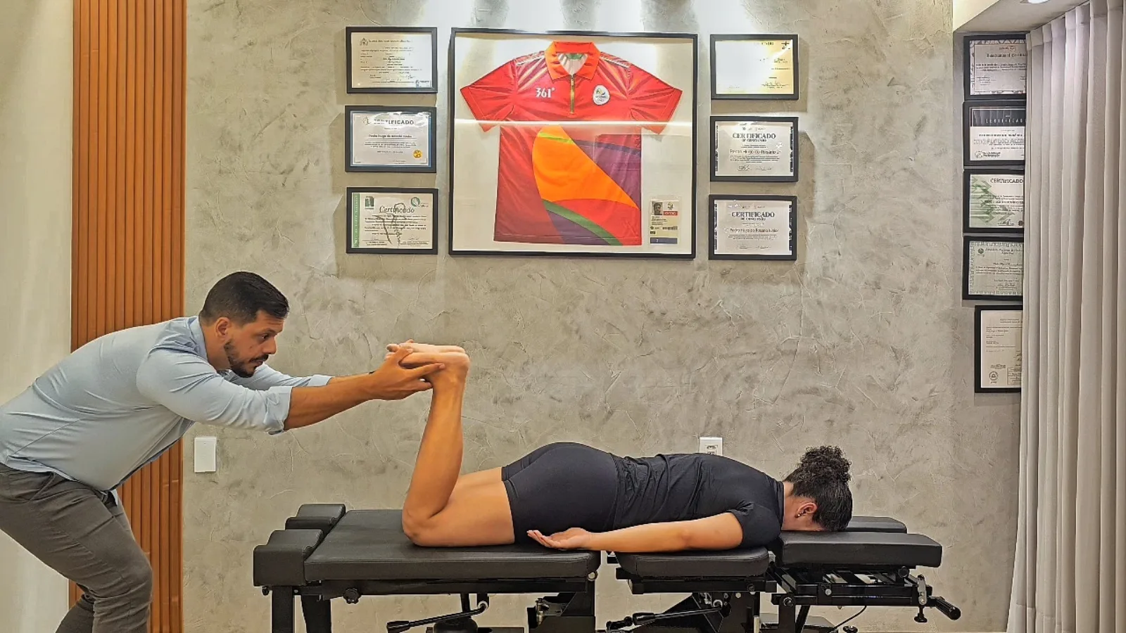 Dr. Pedro Rosário conduzindo teste de mobilidade em paciente, com certificados e camisa olímpica ao fundo